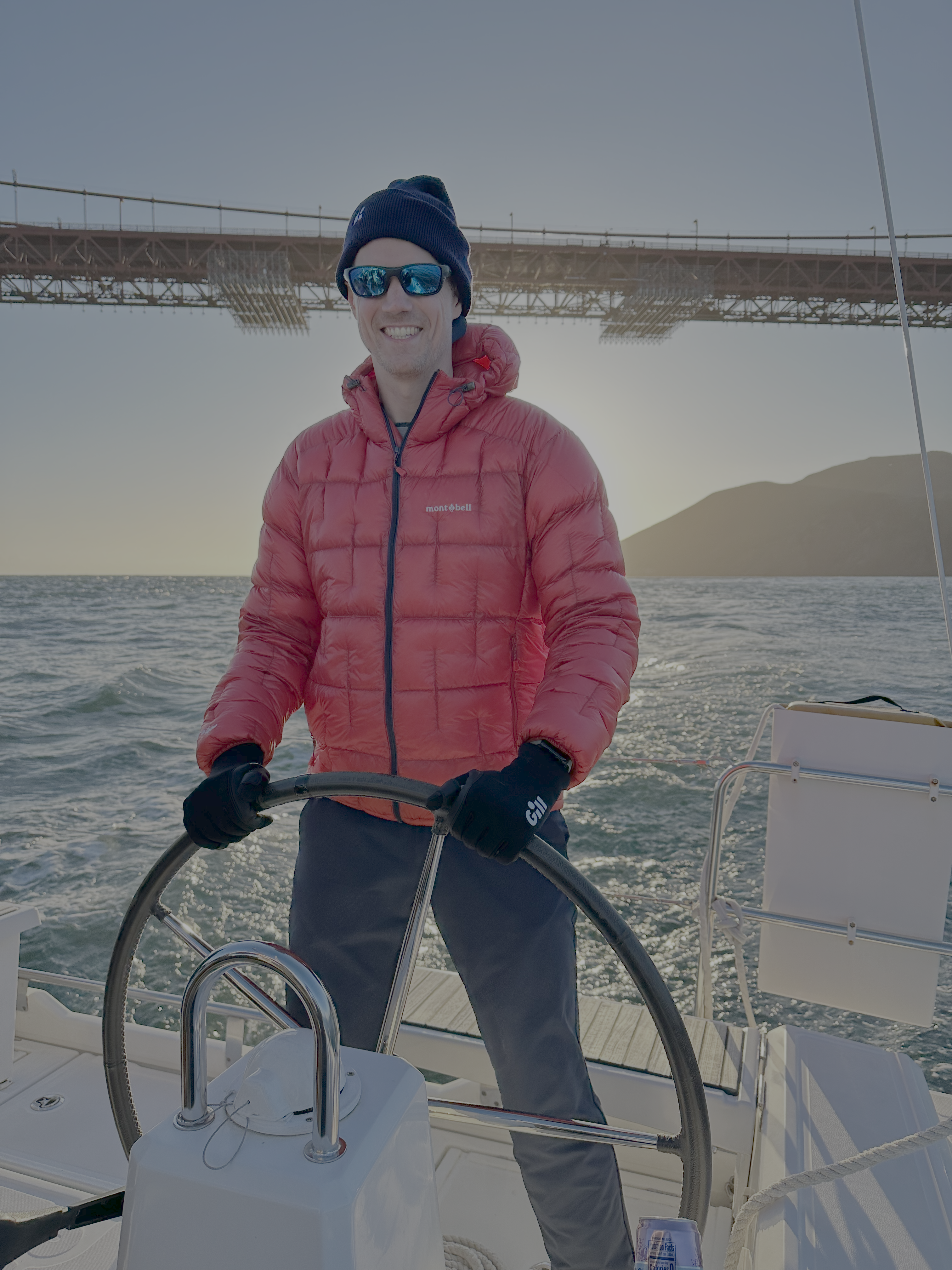 A man smiling with sunglasses on in a red puffy coat, grips the helm of a sailboat with the Golden Gate Bridge behind him