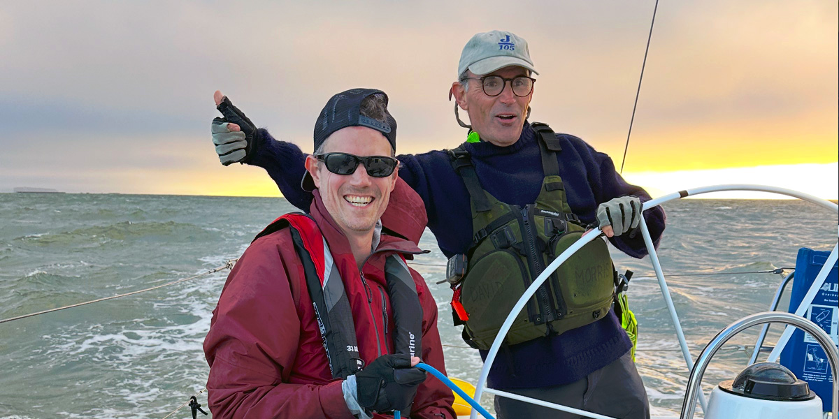 Two men on the back of a racing sailboat, smiling, with an orange and yellow sunset behind them.